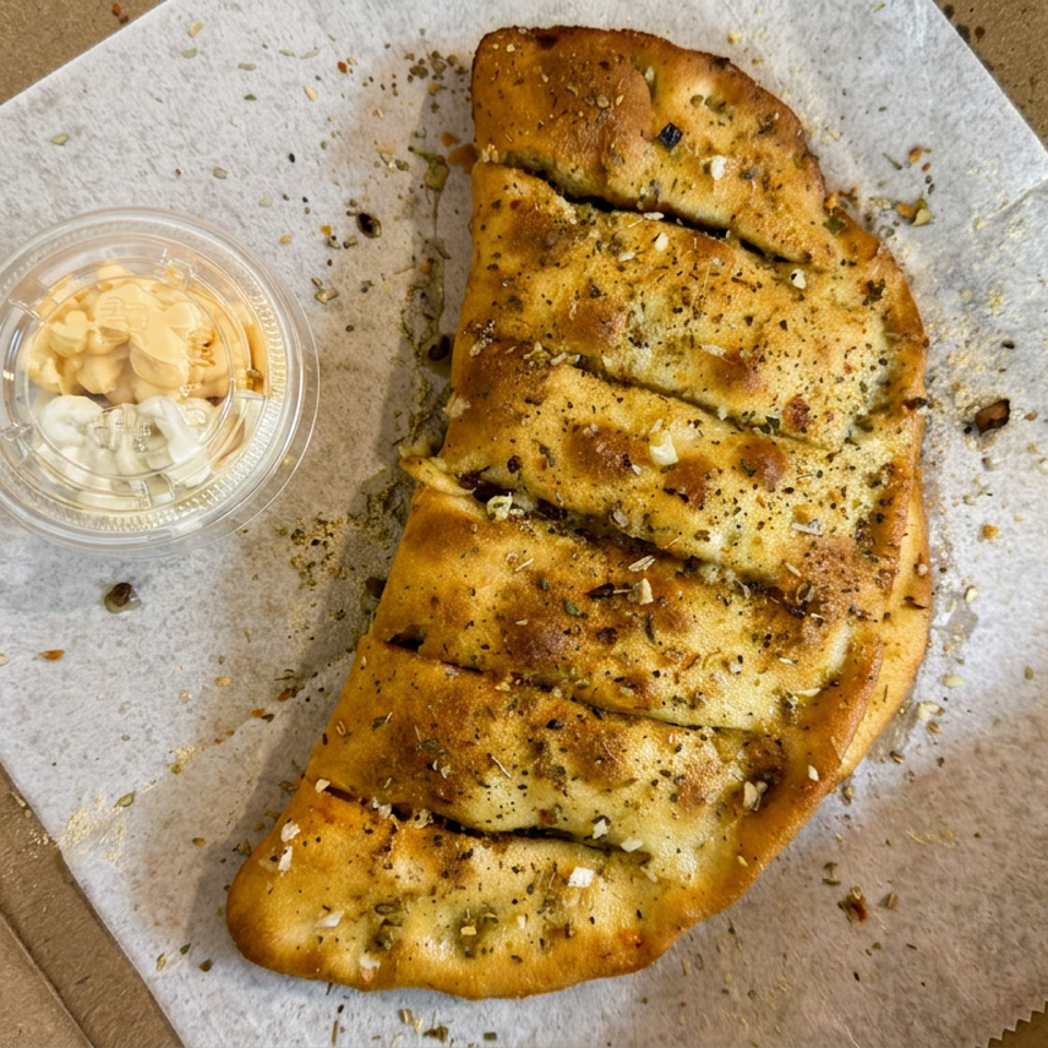 Sliced garlic-and-herb focaccia served in a cardboard box with a small cup of creamy dipping sauce.